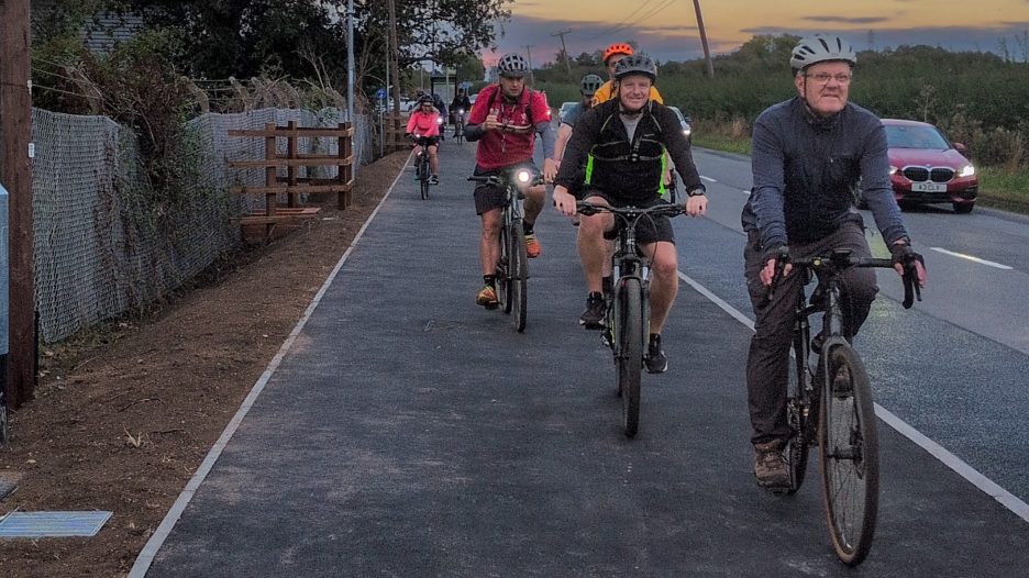 A group of cyclists helmeted and riding along a wide, newly paved shared pedestrian and cycle path next to a road at dusk.
